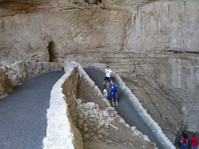 [The switchback path is paved with grey asphalt and lines on both sides with white-tan adobe style stone walls and is completely under the edge of a huge stone overhang. There are kids on the far switchback.]